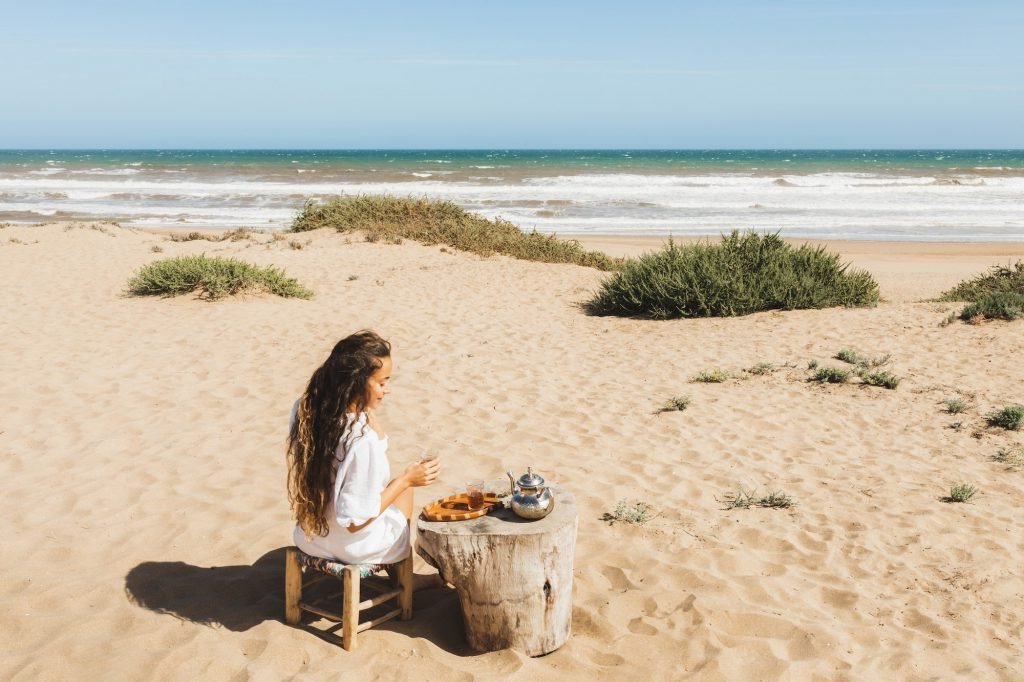 Young happy woman enjoying traditional moroccan mint tea outdoors on ocean sand beach in Morocco